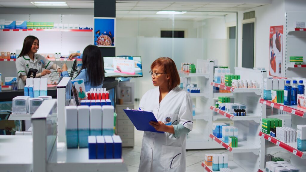 Pharmacist reviewing notes on a clipboard while a coworker helps a customer at checkout